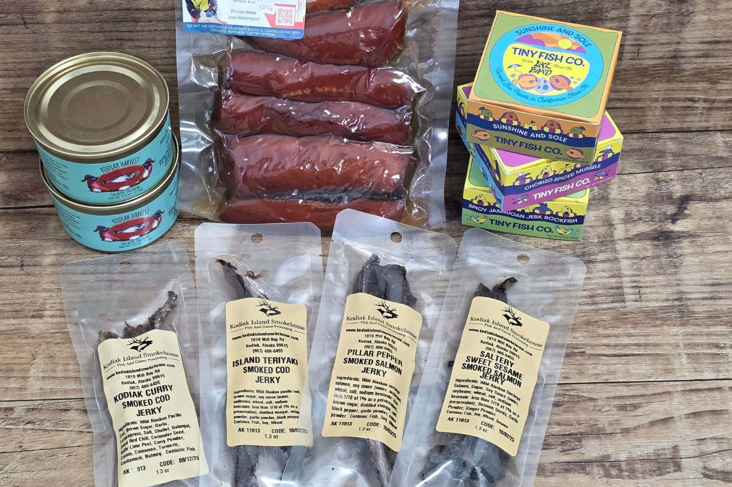 Assorted Kodiak Island Smokehouse seafood products including smoked fish jerky, sausage links, canned fish, and packaged items arranged on a rustic wooden surface
