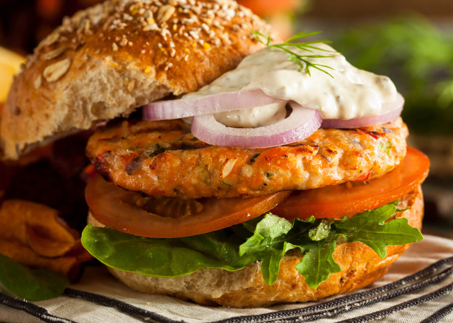 Salmon burger with lettuce, tomato, and onion on a bun with chips and greens in the background.