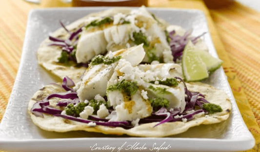 Grilled Alaska halibut tucked into corn tortillas and topped with bright green cilantro-parsley pesto, served with lime wedges and a rustic garnish on a ceramic plate.