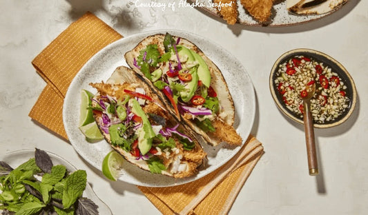 Coconut-crusted Alaska rockfish tacos with cabbage slaw, avocado slices, fresh herbs, and a drizzle of sweet-and-sour Nuoc Cham sauce, served with lime wedges on a rustic plate.