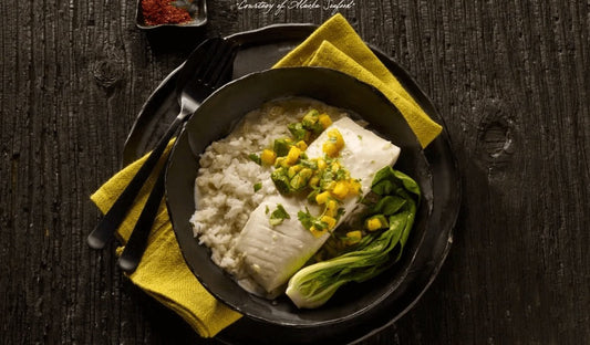 Alaska halibut fillet poached in coconut milk, plated over jasmine rice and topped with mango-avocado salsa, garnished with fresh herbs on a warm-toned plate.