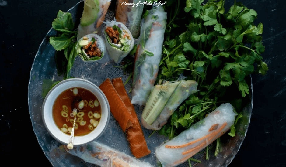 Fresh rolls filled with Alaska smoked salmon, crisp vegetables, basil on a plate with dipping sauce and fresh herbs.