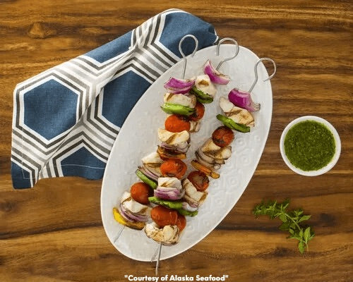 Grilled Alaska rockfish kebabs with charred edges, served with green chimichurri sauce, garnished with lemon wedges and fresh herbs, served on a white platter.
