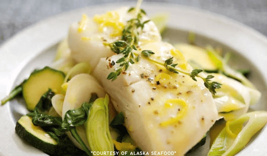 Alaska halibut fillet broiled and topped with lemon-thyme butter sauce, plated with sautéed spinach, zucchini, and leeks on a rustic ceramic plate.