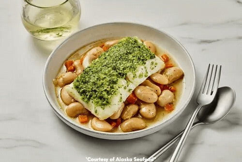 Golden herb-crusted Alaska halibut fillet resting on a bed of white bean cassoulet made with carrots, onion, garlic, and thyme, garnished with fresh herbs on a ceramic plate.