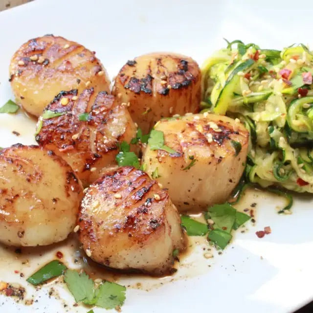 Golden-browned scallops with an Asian-style glaze served on a bed of sautéed zucchini noodles, garnished with chopped cilantro and toasted sesame seeds.
