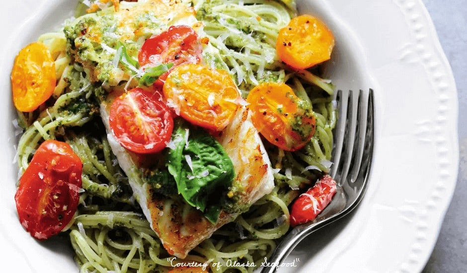 Pan-seared Alaska halibut fillet resting on a bed of pasta tossed in pistachio pesto, topped with roasted cherry tomatoes and garnished with fresh herbs on a ceramic plate.