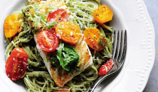Pan-seared Alaska halibut fillet resting on a bed of pasta tossed in pistachio pesto, topped with roasted cherry tomatoes and garnished with fresh herbs on a ceramic plate.