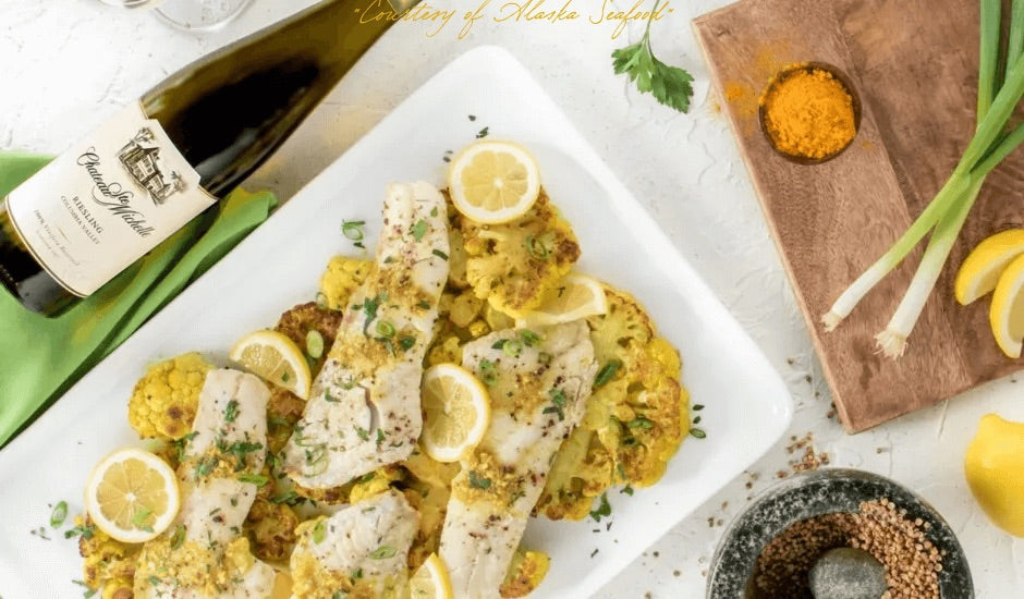 Seared Alaska rockfish fillets and golden roasted cauliflower steaks drizzled with turmeric curry butter, garnished with lemon wedges, scallions, and fresh herbs.