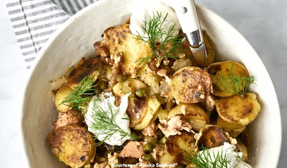 Ceramic bowl of Alaska salmon and potato breakfast hash with bacon, onions, and capers, topped with a dollop of dill-horseradish cream and garnished with fresh dill.