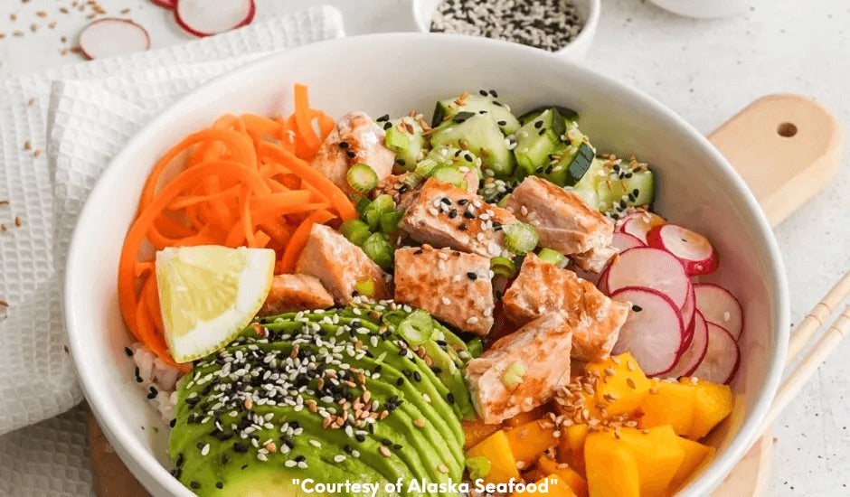 Bowl with cooked wild Alaska salmon, avocado slices, mango, cucumber, radish, and carrot over sushi rice, topped with sesame-teriyaki sauce and sesame seeds.