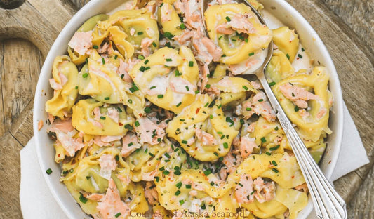 Bowl of cheese tortellini tossed in creamy garlic sauce with chunks of wild Alaska salmon, garnished with parsley and served on a rustic plate.