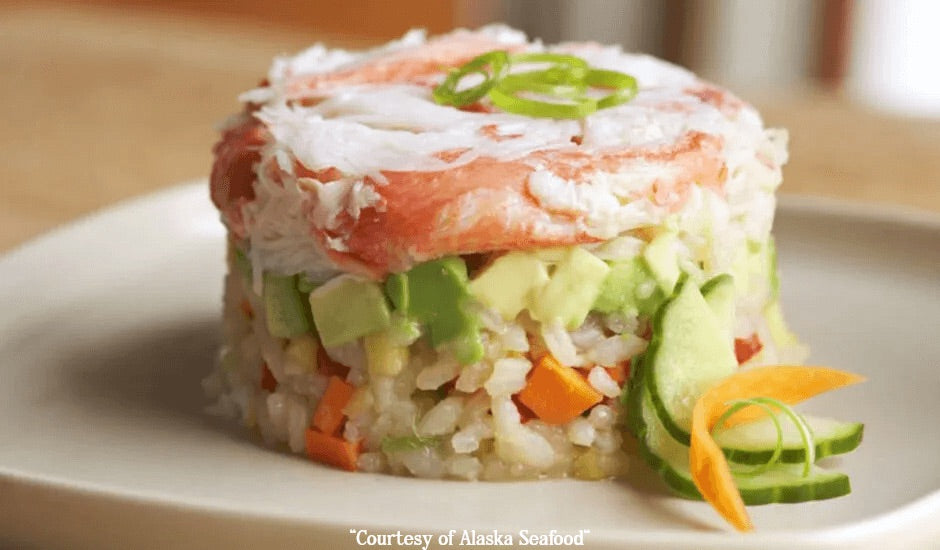 A layered sushi salad featuring Alaska snow crab, seasoned rice, sliced vegetables, and avocado, arranged on a white plate with sliced cucumber garnish.