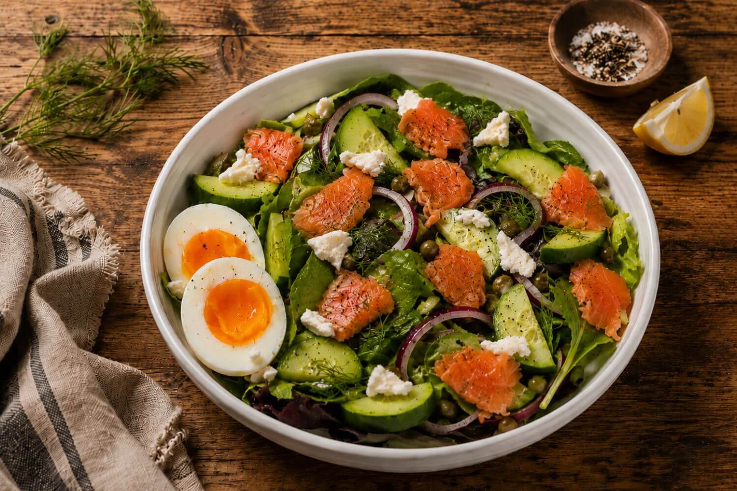Fresh gravlax salad with spring greens, sliced cucumber, pickled red onions, soft-boiled egg, capers, crumbled goat cheese, and lemon-caper dressing in a rustic bowl