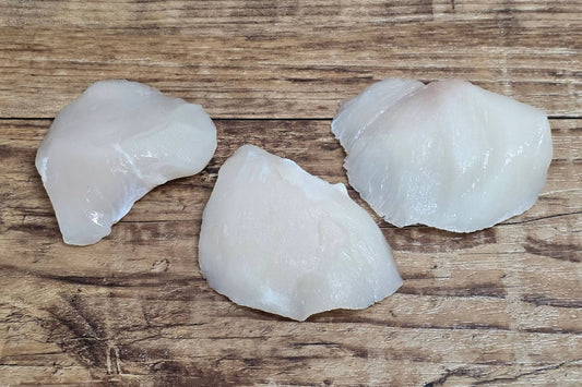 Three raw Wild Alaskan halibut cheeks displayed on a rustic wooden background, showing their natural ivory color, smooth texture, and rounded cheek shape.