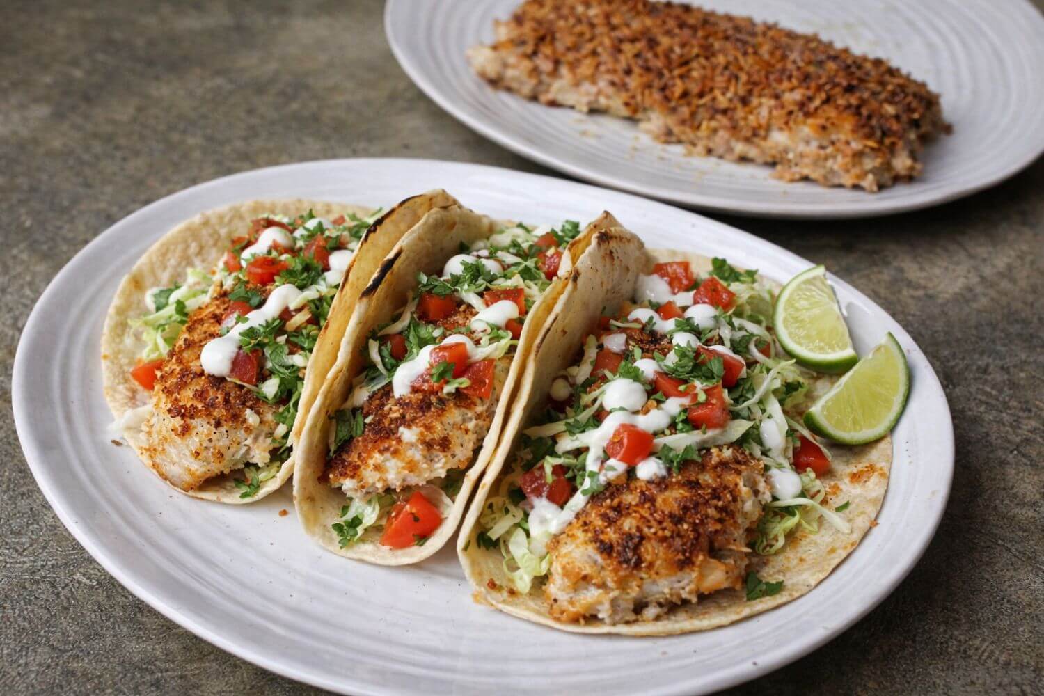Three fish tacos on a white plate with a side of lime wedges on a gray surface.