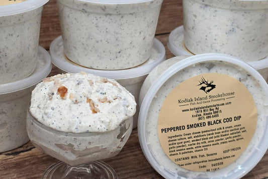 Containers of creamy smoked black cod dip with visible black pepper and sesame seeds on a rustic wood surface.