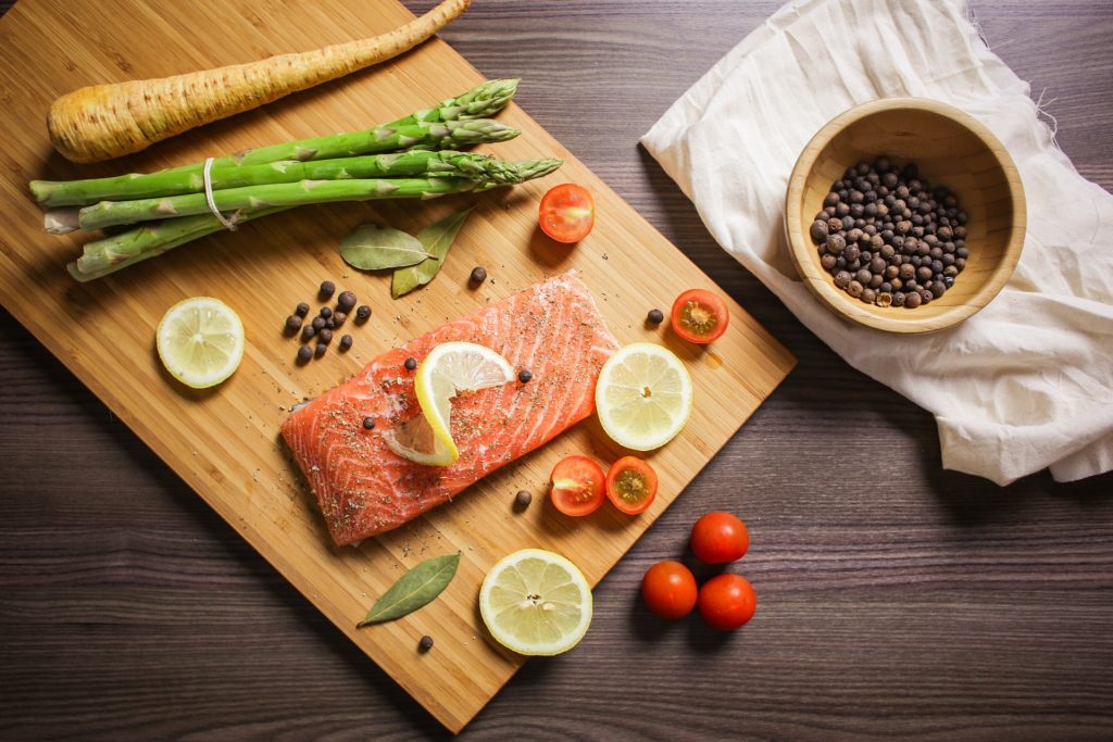 Raw salmon fillet with lemon slices on a wooden board with asparagus, tomatoes, and black peppercorns