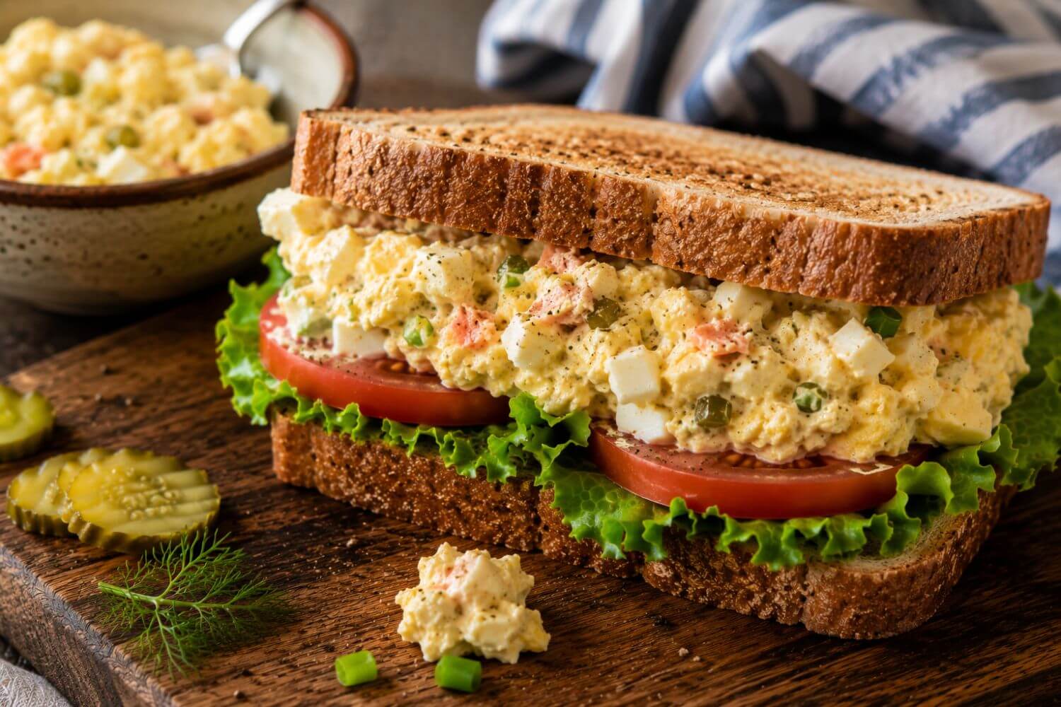 Smoked salmon deviled egg salad sandwich with tomatoes and lettuce on a wood cutting board.