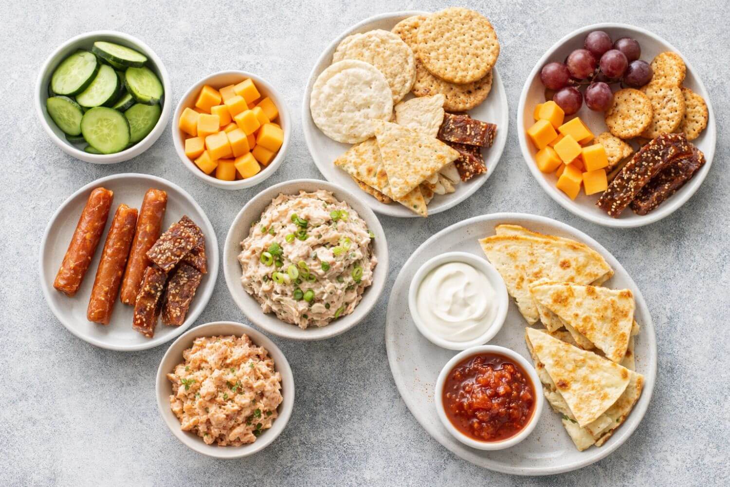 Assorted snacks including crackers, cheese, fruits, and dips, and jerky on a gray surface