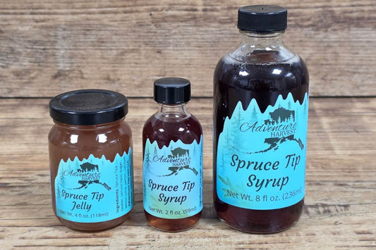 Wild Alaskan spruce tip jelly and spruce tip syrup from Adventure Harvest displayed in glass containers on a rustic wood surface.