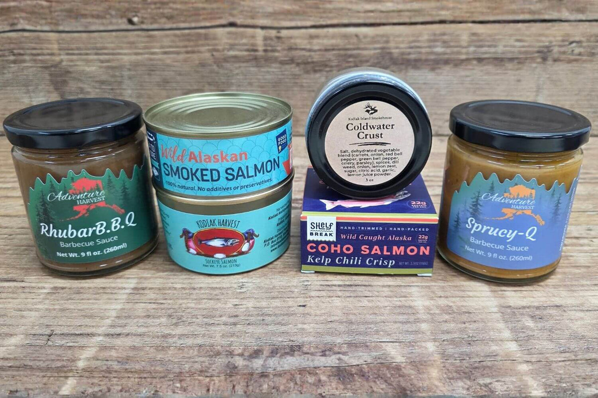 Assortment of Alaska-made smoked salmon, salmon jerky, Coldwater Crust dip, and barbecue sauces arranged on rustic wood.