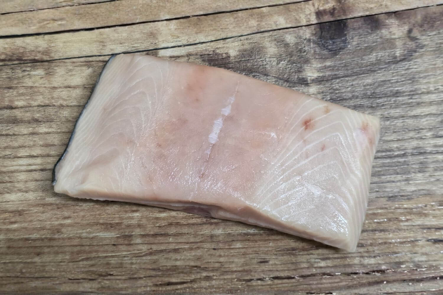 Single raw Wild Alaskan white king salmon fillet portion displayed on a rustic wooden background, showing its pale ivory flesh, subtle marbling, and natural sheen.